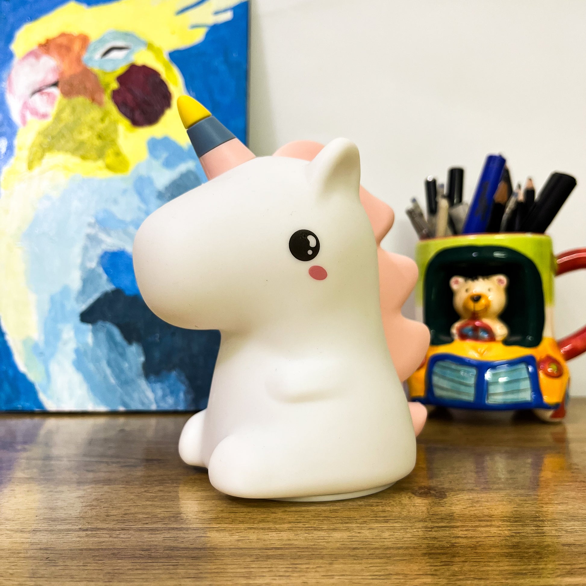 Unicorn-shaped night light on a desk with a colorful painting and pencil holder in the background.