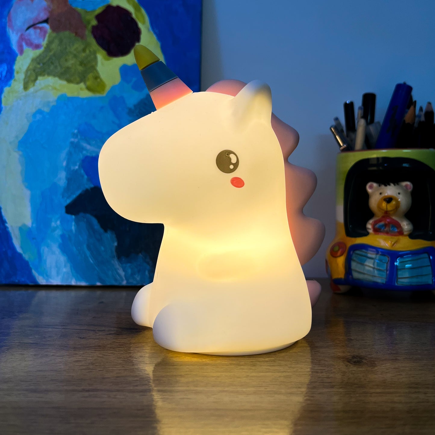 Unicorn-shaped night light on a wooden surface with a colorful painting and stationery items in the background.